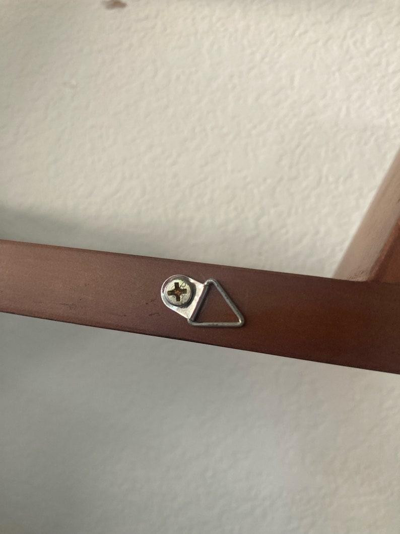 Op de afbeelding: Een close-up van een bruin houten frame met een zilveren driehoekige ophanghaak bevestigd met een schroef. De haak is ontworpen om een ​​foto of ander object aan een muur te hangen.