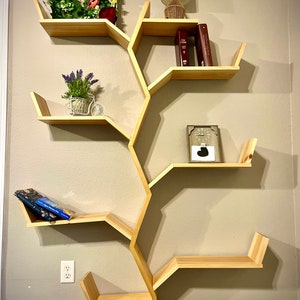 Op de afbeelding: Een houten boomvormig boekenrek met vijf planken. De planken zijn zeshoekig en zijn in een boomvormig patroon gerangschikt. Het boekenrek is versierd met een kleine plant in een pot, een lamp en boeken.