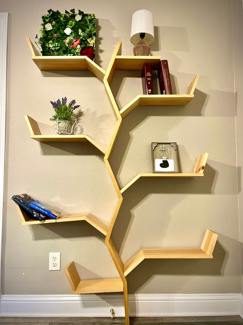 Op de afbeelding: Een houten boomvormig boekenrek met vijf planken. De planken zijn zeshoekig en zijn in een boomvormig patroon gerangschikt. Het boekenrek is versierd met een kleine plant in een pot, een lamp en boeken.