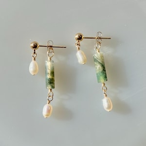 Puede incluir: Un par de pendientes de oro con detalles de ágata verde y perla. Los pendientes presentan una piedra de ágata verde rectangular larga con un alambre de oro envuelto alrededor. Una pequeña perla cuelga de la parte inferior de la piedra de ágata. Los pendientes están unidos a un poste de oro con una pequeña bola de oro en la parte superior.