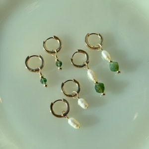 May include: A set of five gold hoop earrings with dangling charms. Two earrings feature a single white pearl charm, while the other three feature a green gemstone charm.