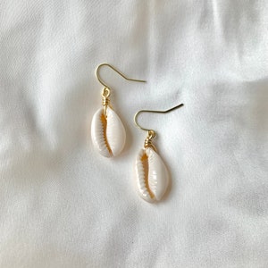 Könnte beinhalten: Ein Paar goldene Ohrringe mit weißen Kaurischnecken. Die Muscheln sind mit Draht an den goldenen Haken befestigt.