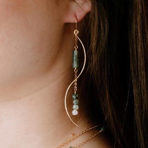 May include: A gold-toned dangle earring featuring a curved metal wire, a delicate chain, and a series of green and white beads. The earring includes a cylindrical green bead at the top and three smaller spherical beads in varying shades.
