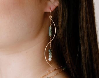 May include: A gold-toned dangle earring featuring a curved metal wire, a delicate chain, and a series of green and white beads. The earring includes a cylindrical green bead at the top and three smaller spherical beads in varying shades.
