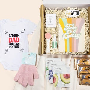 May include: A gift box with a white baby onesie that says "C'mon Dad You Can Do This". The box contains a notebook with "Take it Easy" text, skincare products, a pink face scrubber, and gold foil packets.