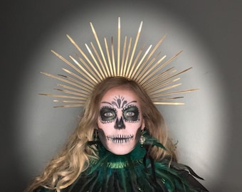 May include: A person with Day of the Dead makeup, including a skull face design, wearing a gold halo and a dark green feather collar. The person has long blonde hair and is looking directly at the camera.