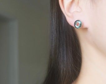 May include: A pair of small, round, teal-colored earrings with a silver setting. The earrings are worn in the earlobe of a person with long, dark hair.