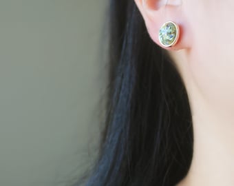 May include: A pair of oval-shaped earrings with a green and gold design. The earrings are made of metal and have a shiny finish.