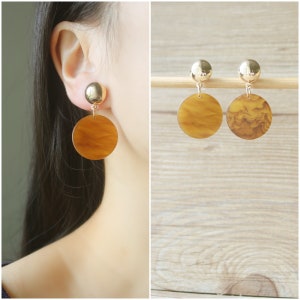 May include: Gold-tone stud earrings with a large, circular, amber-colored pendant. The earrings have a marbled pattern and are shown on a person's ear and also displayed on a wooden dowel against a wood-grain background.