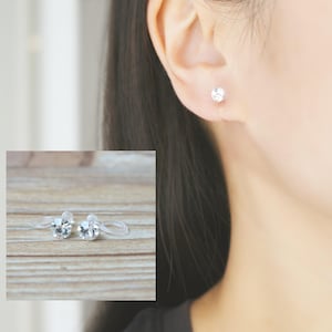 May include: A pair of clear, plastic, non-pierced earrings with a small, clear rhinestone in the centre. The earrings are shown on a wooden surface and in a person's ear.