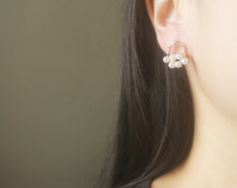 May include: A gold-toned earring featuring a cluster of small, white pearls. The earring has a delicate, openwork design, with the pearls suspended from a curved, circular frame. The earring is shown on a person's ear, with the focus on the jewelry.
