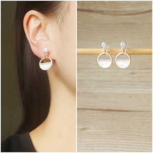 May include: A pair of gold-tone earrings with a white circle design and a row of clear rhinestones. The earrings are on a wooden surface.