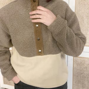 Peut inclure: Un pull en polaire marron et beige avec une patte de boutonnage en velours côtelé. Le pull a un col montant et des manches longues.