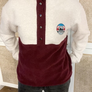 Peut inclure: Un pull en polaire blanc et bordeaux avec une fermeture à boutons pression. Le pull a un patch sur la poitrine gauche avec une scène de montagne et le texte "Mountain Explorer".