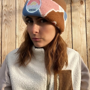 Pode incluir: Um gorro de lã polar castanho com um patch colorido com um design de montanha e sol. O gorro tem um padrão rosa, azul e branco. O texto "BINOUX" é visível no patch. A pessoa está a usar um casaco de lã polar branco.
