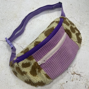 Puede incluir: Una riñonera morada y marrón con cierre de cremallera. La riñonera está hecha de un material suave y peludo y tiene una correa morada.