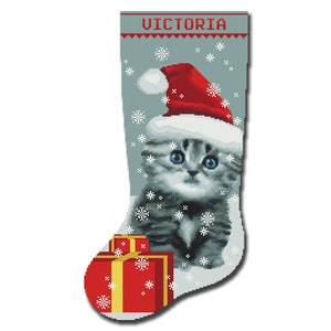 Può includere: Un calzino di Natale grigio e bianco con il nome "VICTORIA" ricamato in alto. Un gattino grigio e bianco con un cappello da Babbo Natale rosso è seduto dentro il calzino con una scatola regalo rossa e dorata.