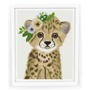 May include: A framed cross-stitch art piece featuring a cheetah cub wearing a floral crown. The cheetah has tan fur with dark spots, large brown eyes, and a white muzzle. The floral crown includes blue and cream-colored flowers with green leaves.