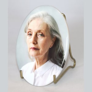 May include: A white oval ceramic plate with a portrait of a woman with grey hair. The plate is on a gold metal stand.