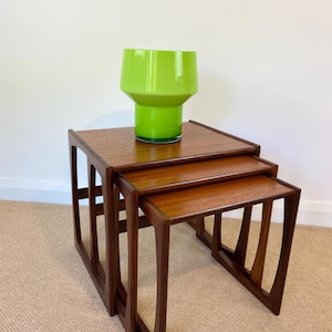 May include: A set of three nested wooden tables with a rich brown finish and angular legs, arranged in a tiered formation. A bright lime green glass vase with a wide, flared top and a cylindrical base sits on the largest table surface.