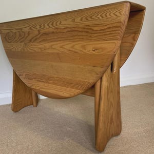 May include: A light brown wooden drop-leaf table with a prominent natural wood grain pattern. The table features a rectangular top with two rounded, hinged side leaves that hang vertically, supported by a sturdy base with curved legs.