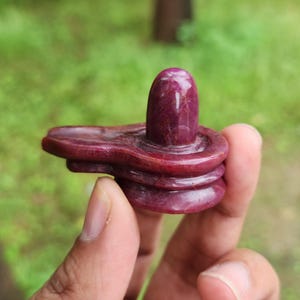 Może przedstawiać: Rzeźbiona, ciemnoczerwona rzeźba z kamienia z zaokrąglonym wierzchołkiem i warstwową podstawą. Rzeźba jest trzymana w dłoni na rozmytym zielonym tle. Kamień ma wypolerowany wygląd.