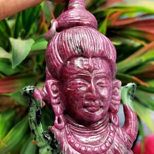 Può includere: Un ciondolo in rubino intagliato che raffigura una divinità con un terzo occhio, che indossa una corona e gioielli elaborati. Il ciondolo è finemente dettagliato e presenta una pietra verde e nera sui lati.