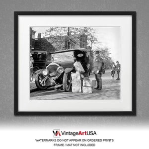 May include: A black and white photograph of a vintage car being unloaded. The car is parked on a street and has a license plate that reads 