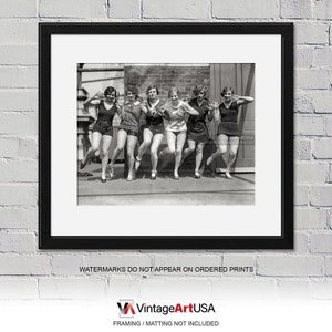 May include: Black and white photograph of seven women in bathing suits posing on a rooftop. The women are all smiling and looking at the camera. They are wearing short skirts and sleeveless tops. The photo is taken from a low angle, and the women are standing in a line.