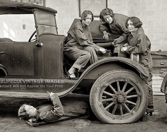May include: Four women in overalls work on a vintage car in a garage. One woman is lying under the car, while the other three are standing around it. The car is a black open-top vehicle with a spare tire on the back.