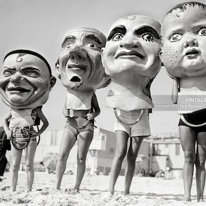 May include: Four people are wearing oversized, comical masks on a beach. The masks are all different and feature exaggerated facial expressions. The people are dressed in swimsuits and are standing in a line.