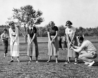 May include: Five women in white and black clothing are being taught how to play golf by a man in a white shirt and khaki trousers. The women are holding golf clubs and are standing on a grassy field. The man is demonstrating the correct stance and swing.
