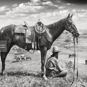 Puede incluir: Una fotograf&iacute;a en blanco y negro de un vaquero sentado en el suelo junto a un caballo. El vaquero lleva un sombrero y una camisa de manga larga. El caballo lleva una silla de montar y una brida. El fondo es un campo con otros caballos en la distancia.