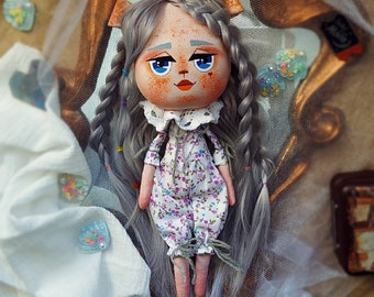 May include: A handmade doll with a cat-like face, gray braided hair, and blue eyes. The doll wears a white floral jumpsuit with a lace collar. The doll has pink feet and arms. A small bunny-shaped embellishment is attached to the doll's head.