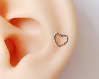 May include: A silver heart-shaped earring with an open center. The earring is shown in a person's ear.