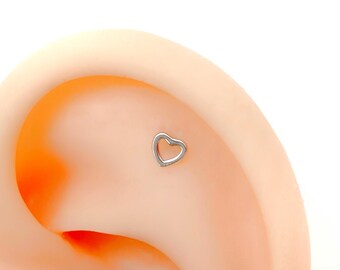 May include: A close-up of a silver heart-shaped earring stud, worn in the cartilage of an ear.