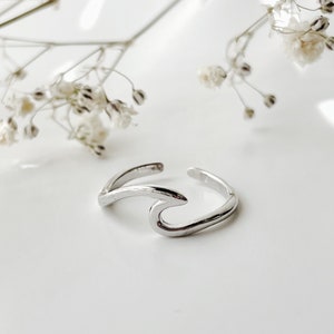 Puede incluir: Un anillo de plata con forma de ola. El anillo está hecho de metal y tiene la forma de una ola. Es un diseño simple y elegante.
