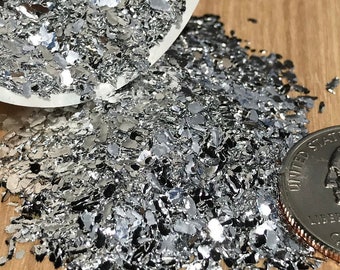 May include: A white ceramic bowl filled with silver glitter, spilling out onto a wooden surface. A US quarter is placed next to the glitter for scale.