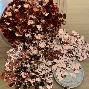 May include: Copper-colored glitter flakes spilling from a clear container. The flakes are scattered on a wooden surface, with a US quarter coin nearby for size comparison. The glitter has a metallic, reflective quality.