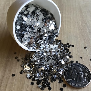 May include: A white container tipped over, spilling silver glitter onto a wooden surface. A US quarter coin is next to the glitter, providing a size reference. The glitter is in the shape of small, flat circles.