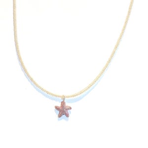 May include: A fine necklace with a small, textured reddish-brown starfish pendant. The chain is a light gold colour, creating a simple yet elegant design. The necklace is displayed against a clean white background.