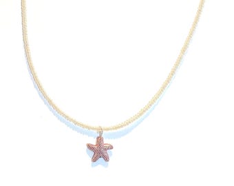Peut inclure: Un collier délicat avec un petit pendentif en forme d'étoile de mer texturée de couleur brun rougeâtre. La chaîne est de couleur or clair, créant un design simple mais élégant. Le collier est présenté sur un fond blanc.
