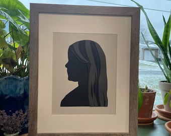 May include: A framed silhouette of a person with long hair, the silhouette is black and the background is a light gray. The frame is a light brown wood.