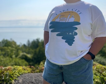 May include: White t-shirt with a graphic of a city skyline, a yellow sun, and blue water. The text "LAKE CITY" is arched above the skyline. The person is wearing blue denim shorts and a black watch.