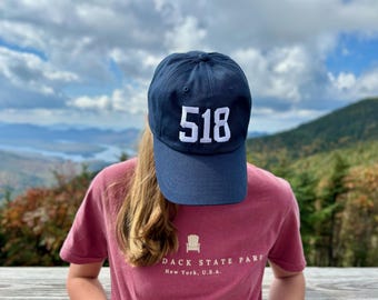 May include: Navy blue baseball cap with the number "518" in white block letters. A person is wearing the cap and a maroon t-shirt with text and a graphic of a chair. The background shows a mountain range and cloudy sky.