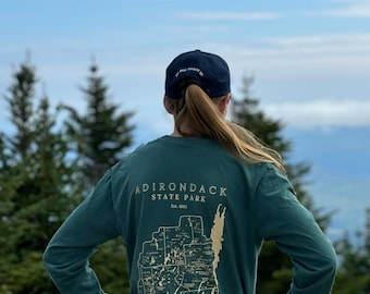 May include: A teal long-sleeve shirt with a map of Adirondack State Park, New York, printed on the back. The text "Adirondack State Park Est. 1892" is printed above the map. The shirt is worn by a person wearing a navy blue baseball cap with the text "The Wild Provides" printed on the back.