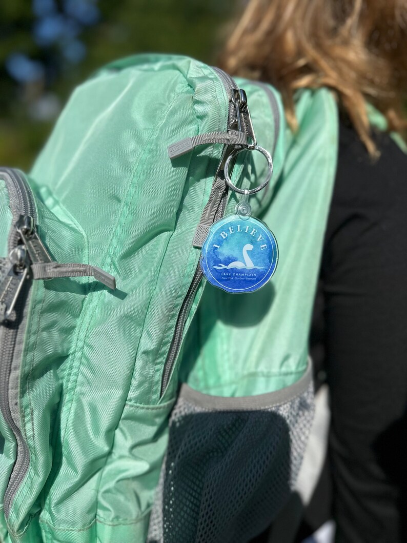 Puede incluir: Una mochila verde claro con un bolsillo con cremallera y un bolsillo de malla. Un llavero redondo con un dise&ntilde;o azul y blanco est&aacute; unido al tirador de la cremallera. El llavero presenta una imagen de dibujos animados de un monstruo marino con el texto "I Believe" y "Lake Champlain" impreso en &eacute;l.