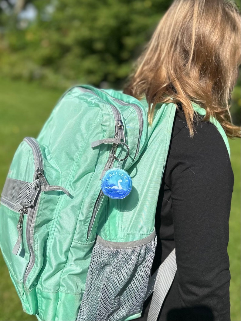 Puede incluir: Una mochila verde claro con un bolsillo con cremallera y un bolsillo de malla. Un llavero con un dije redondo azul y blanco que dice "Creo" y tiene una imagen del monstruo del Lago Ness est&aacute; unido a la mochila.