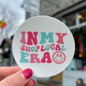 Peut inclure: Un autocollant rond blanc avec le texte "In My Shop Local Era" en rose, bleu et blanc en police rétro. Un smiley rose est à droite du texte.