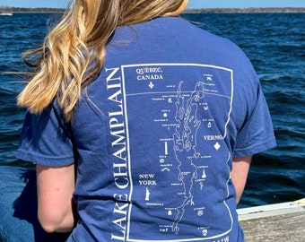May include: A blue t-shirt with a white graphic of a map of Lake Champlain. The map shows the borders of Vermont, New York, Quebec, and Canada. The text "LAKE CHAMPLAIN" is written vertically on the left side of the map. The text "44.5866°N 73.3801°W" is written below the map.